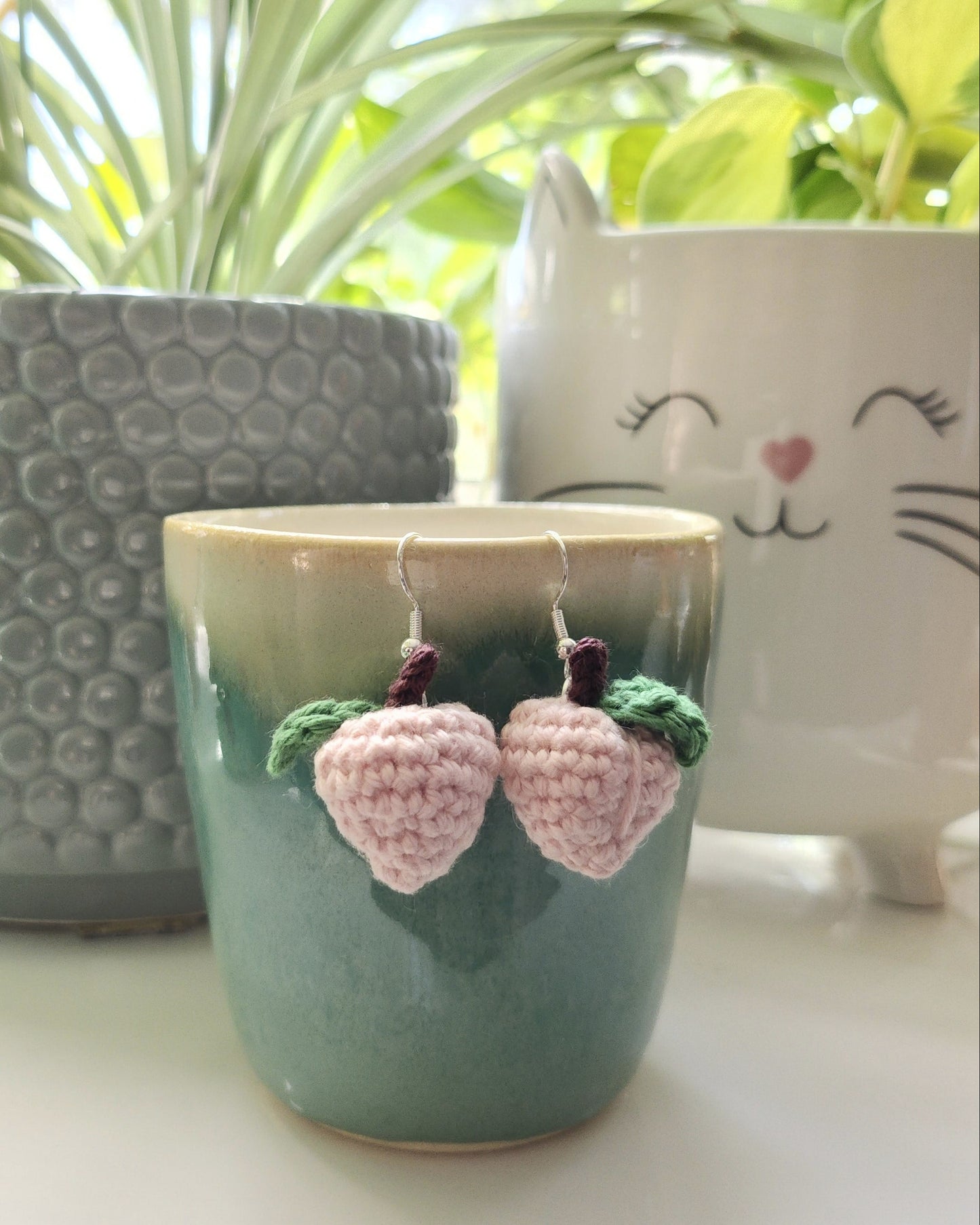 Crochet peach earrings on a handmade cup, placed on a surface with plants in the background.