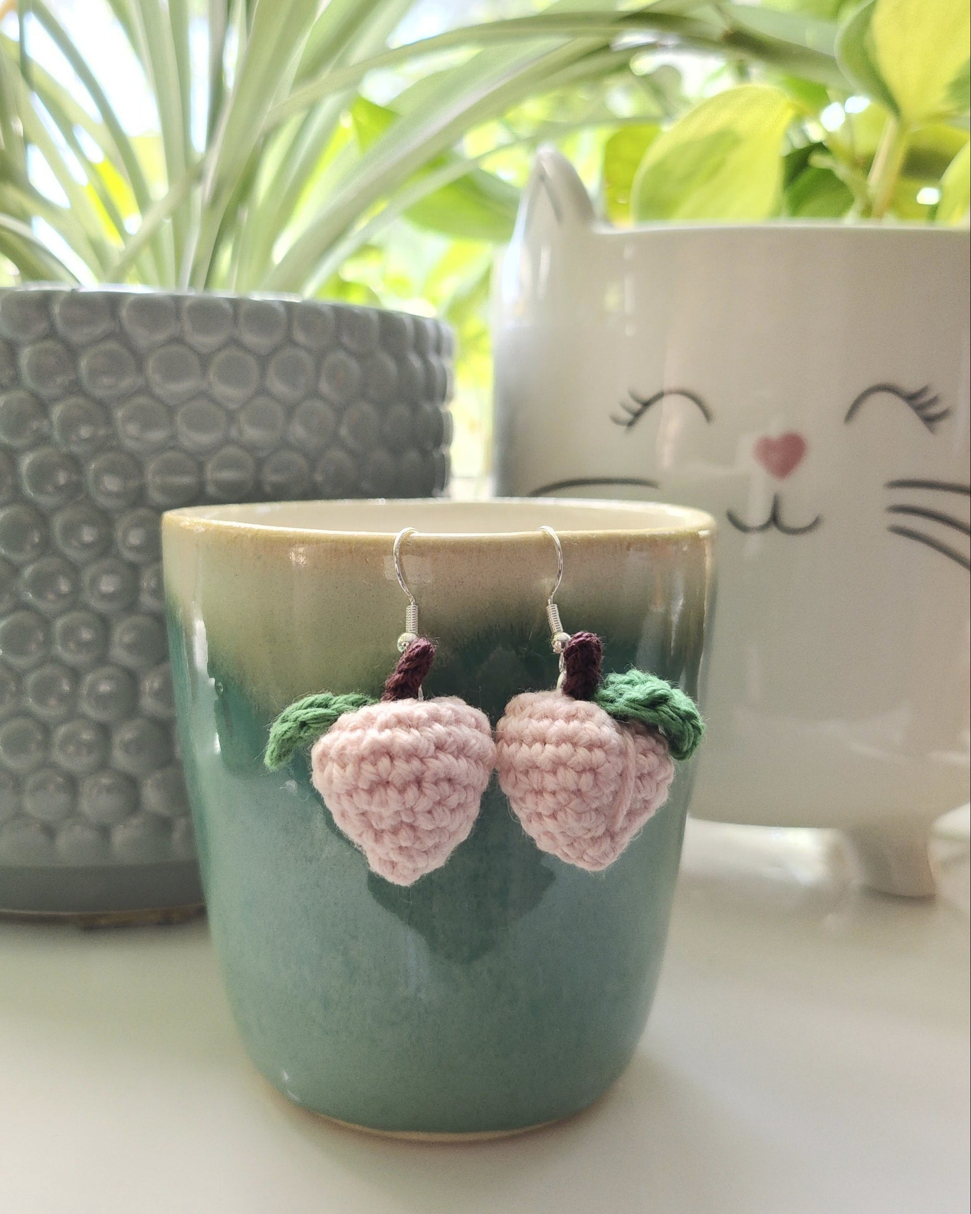 Crochet peach earrings on a handmade cup, placed on a surface with plants in the background.