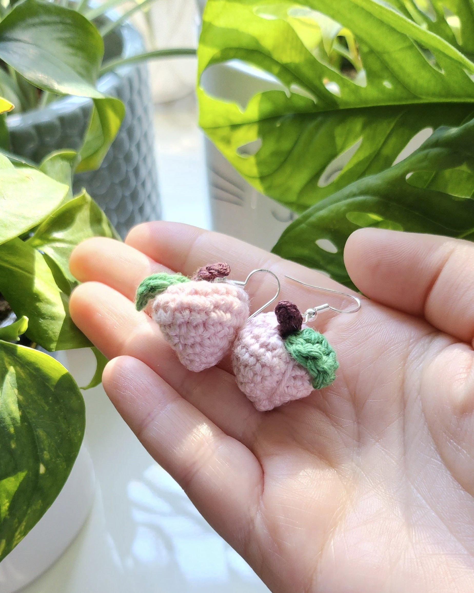 Hand holding two small, colorful peach earrings against a blurred indoor background with plants.