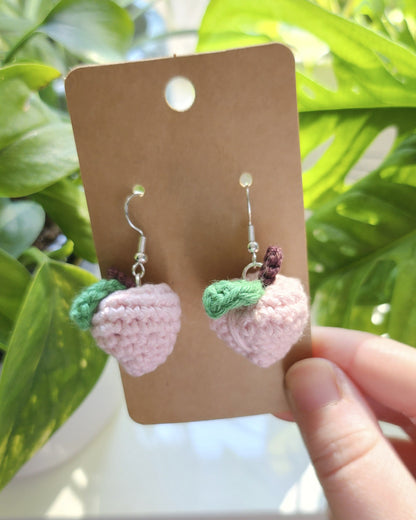 Hand holding a card with pink and green crochet earrings against a blurred plant background