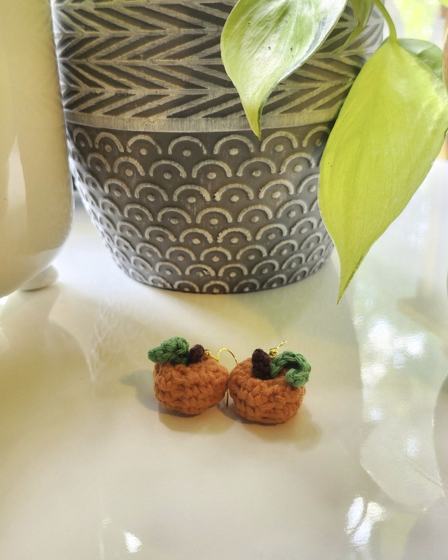 Pair of orange crochet pumpkin earrings on a white surface with a patterned plant pot in the background.