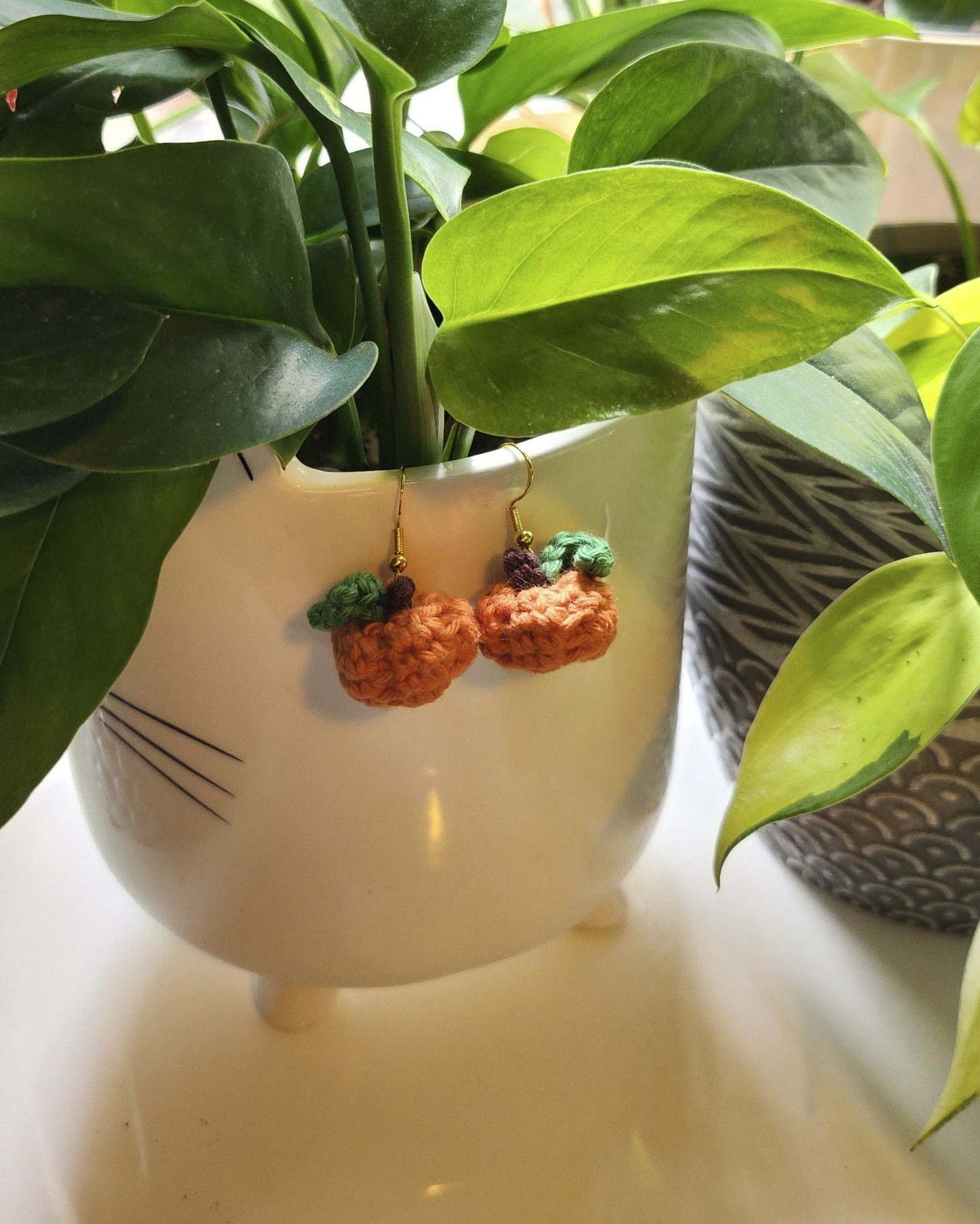 White ceramic planter with green leaves and a small pair of pumpkin earrings inside, surrounded by other plants.