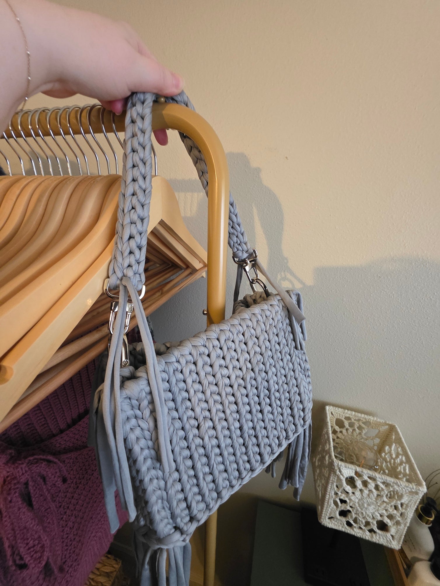 Gray knitted handbag held by a hand with a wooden hanger and decorative items in the background.