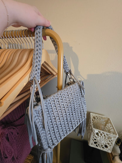 Gray knitted handbag held by a hand with a wooden hanger and decorative items in the background.